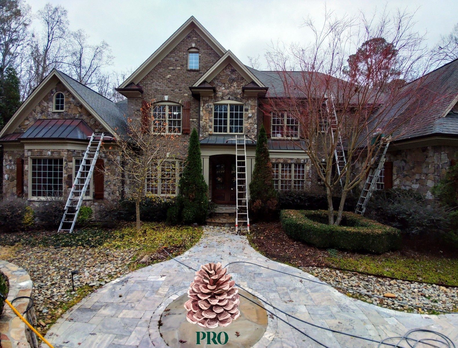 A two-story stone house with ladders against its walls and a decorative pinecone logo on the paved front walkway.