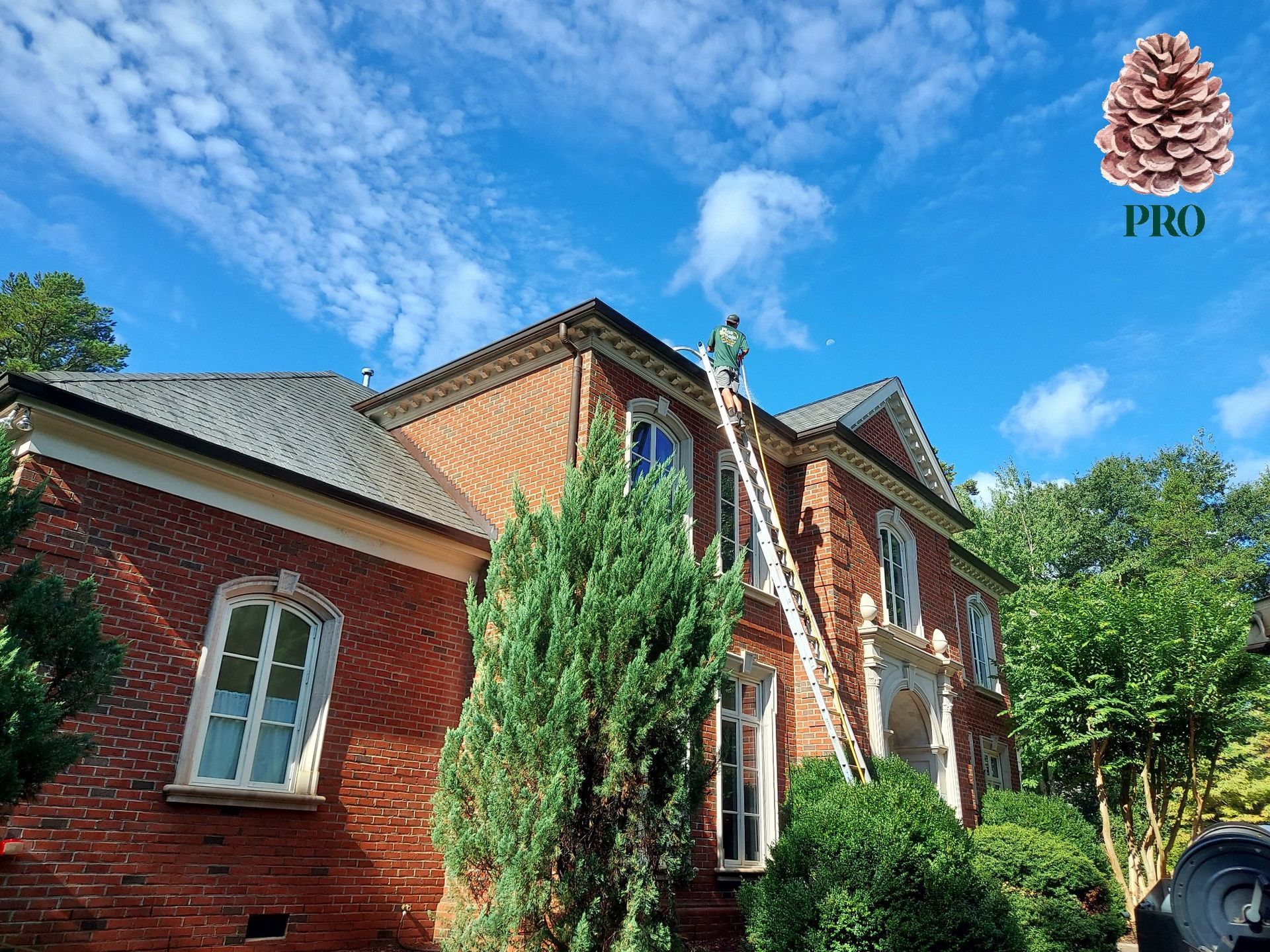A worker stands on a tall ladder cleaning the gutters of a two-story red brick house under a blue, cloudy sky.