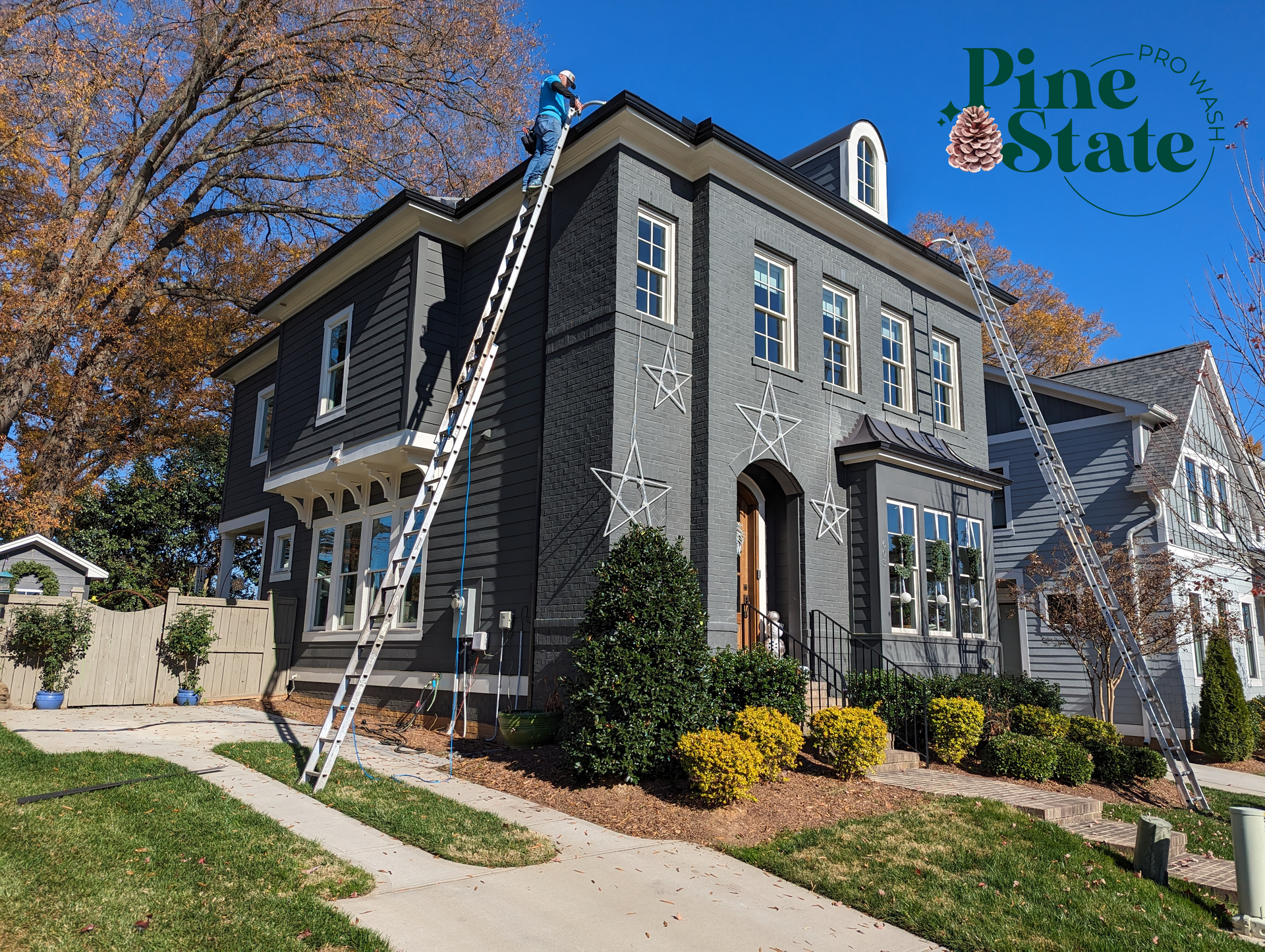 A worker on a ladder installs lights on the exterior of a dark gray two-story house on a sunny day.
