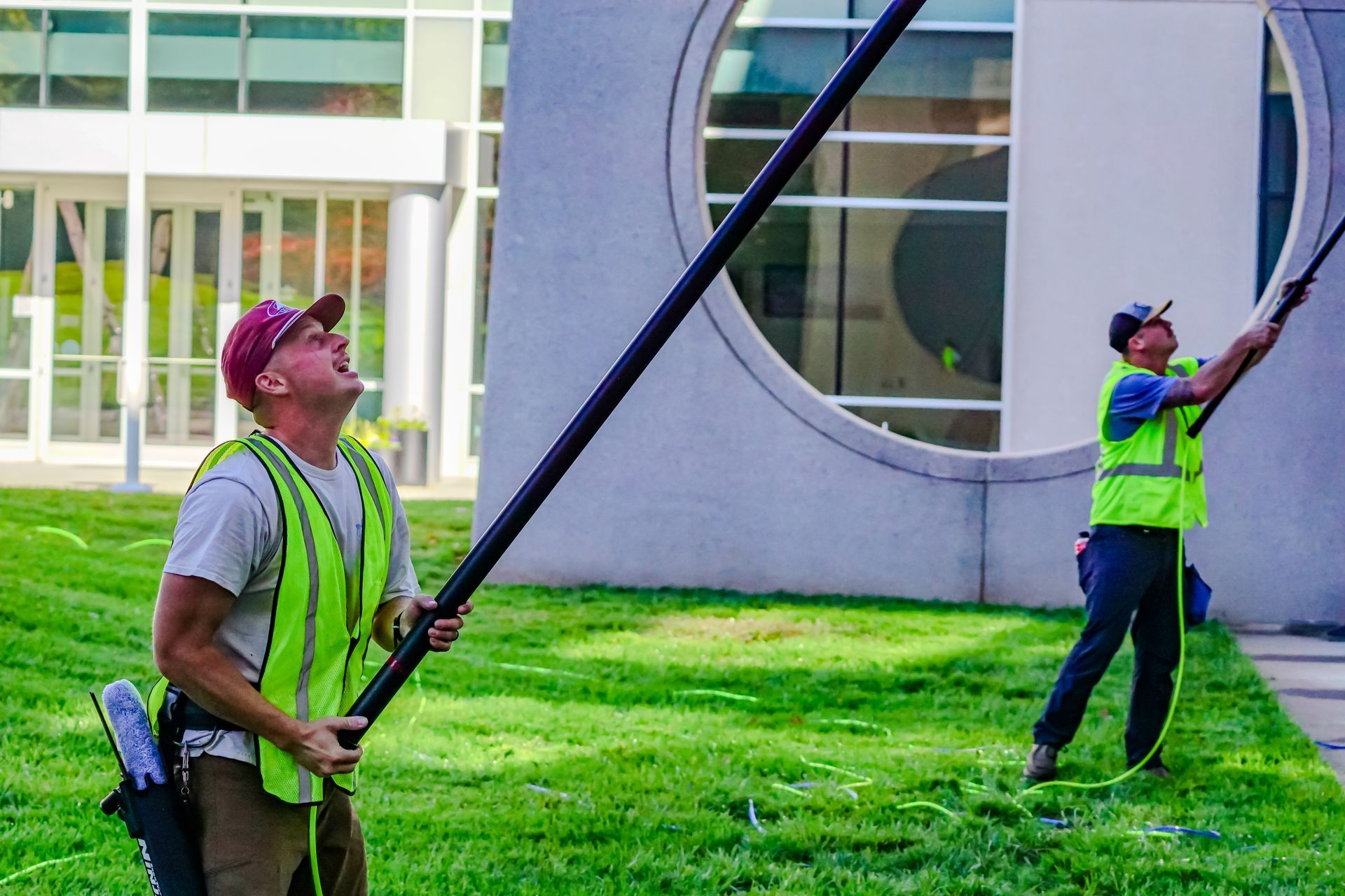 Two workers in neon safety vests use long poles to clean high windows on a building exterior.