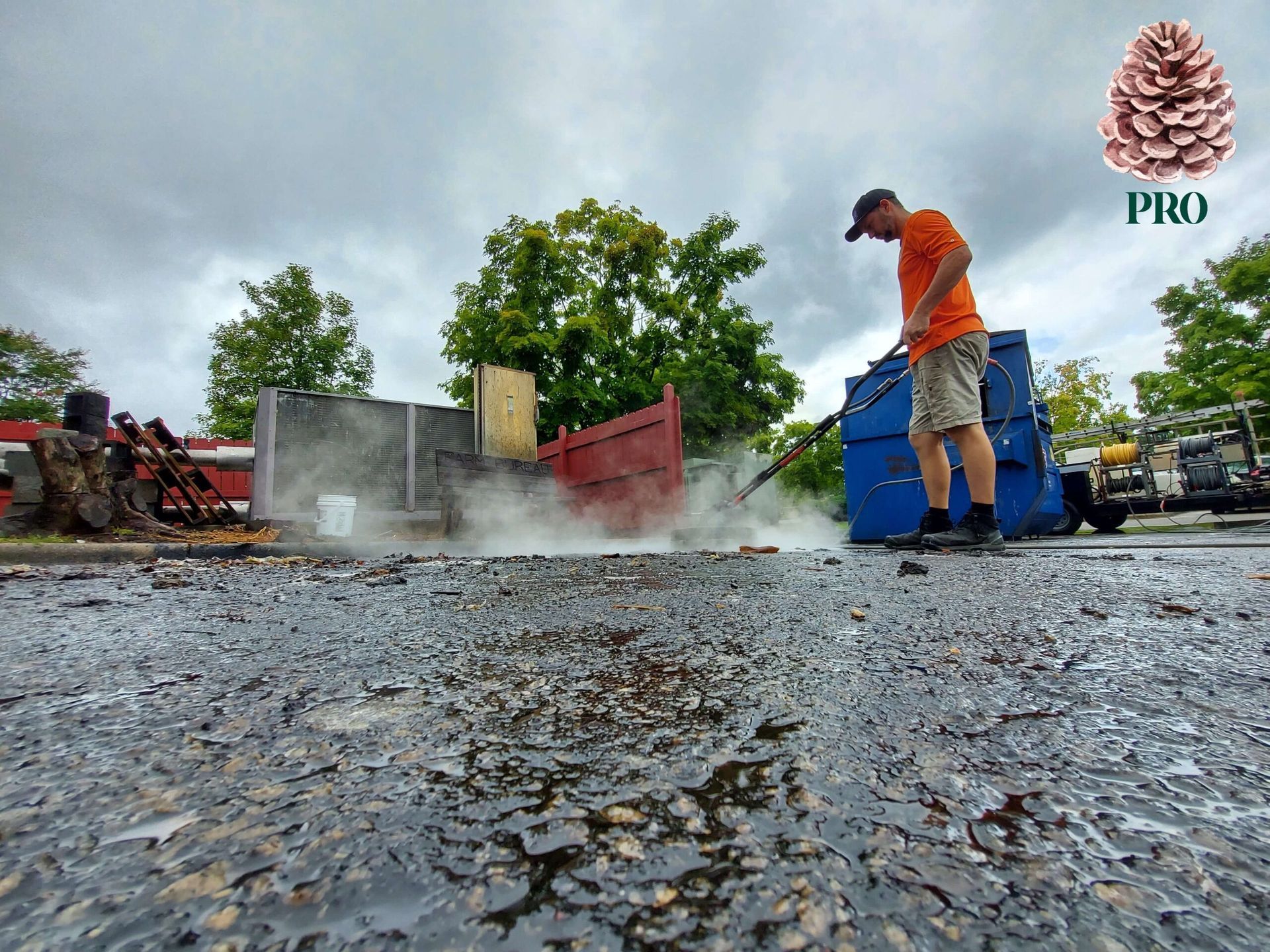 A worker in an orange shirt uses a pressure washer on a paved surface near blue industrial dumpsters.
