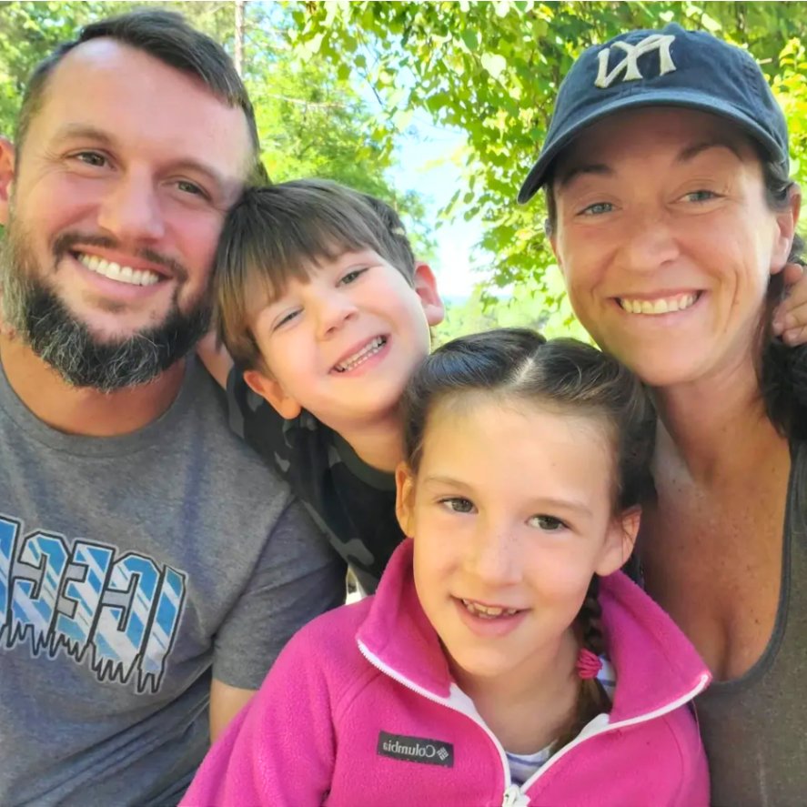 A smiling family of four poses closely together outdoors in front of green trees.
