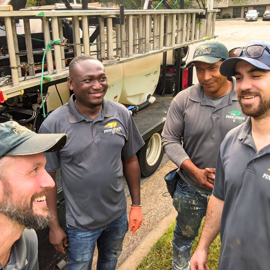 Four smiling workers in company shirts and hats stand together outdoors in front of a work trailer with a ladder rack.