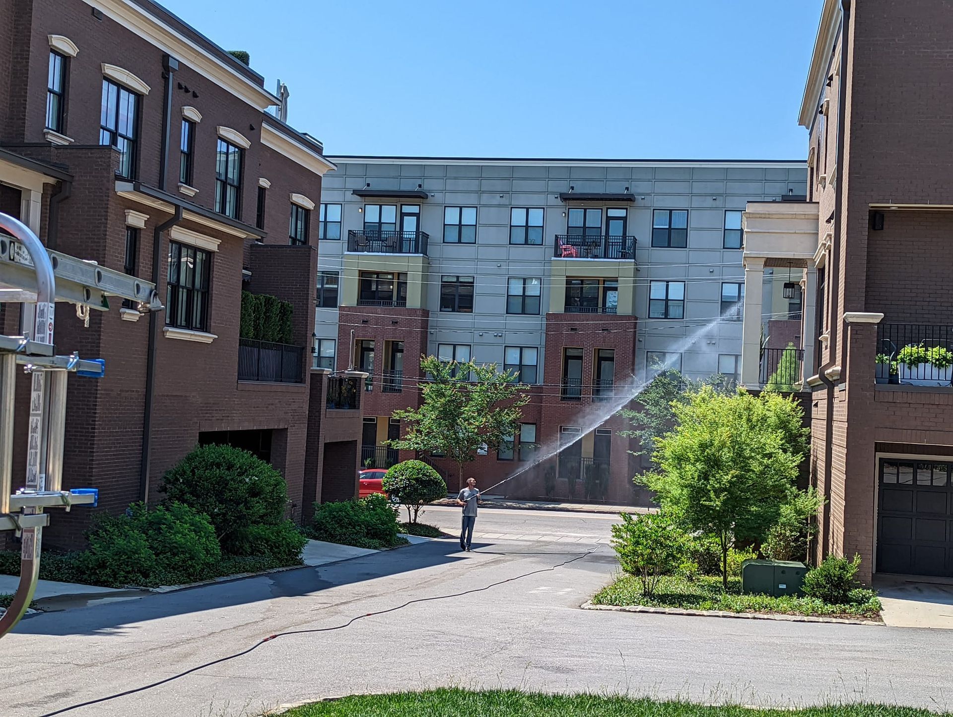 A person sprays water from a hose against the brick exterior of a residential building in an alleyway.