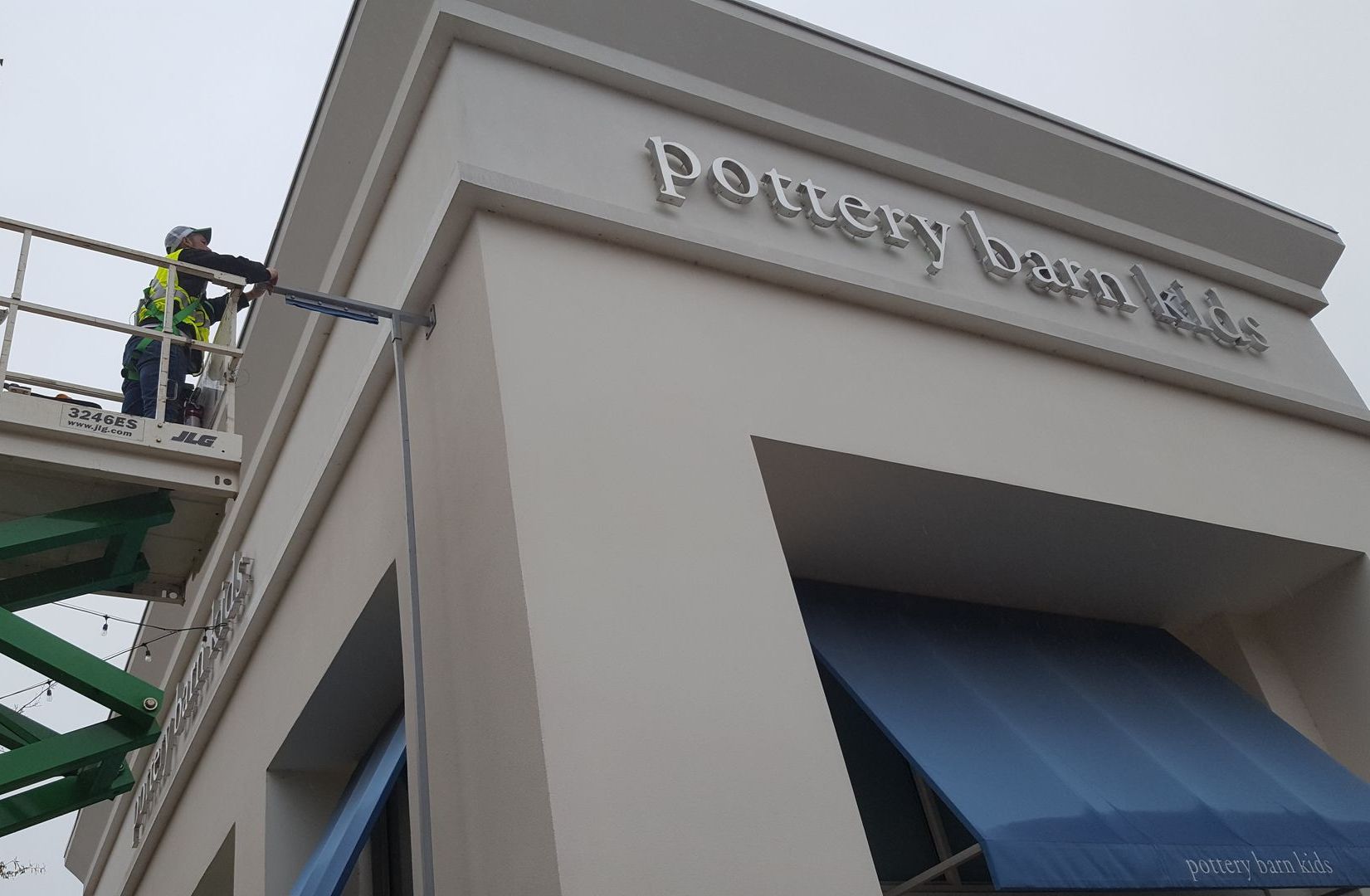 A worker in a safety vest stands on a lift, installing signage on the beige facade of a Pottery Barn Kids store.