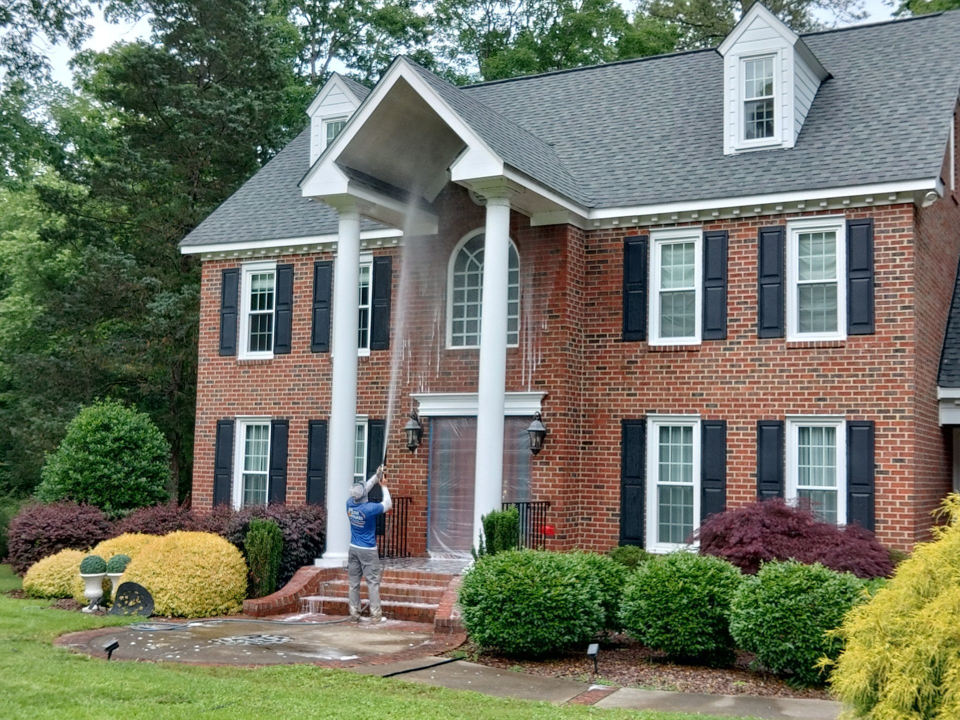 A man is cleaning the front of a large brick house with a hose.