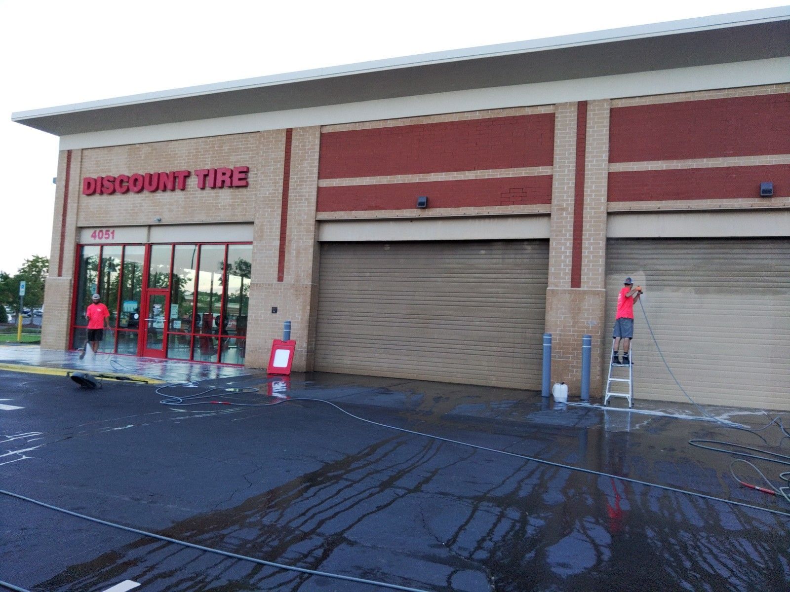 Two workers pressure wash the exterior walls and metal roll-up doors of a Discount Tire store.