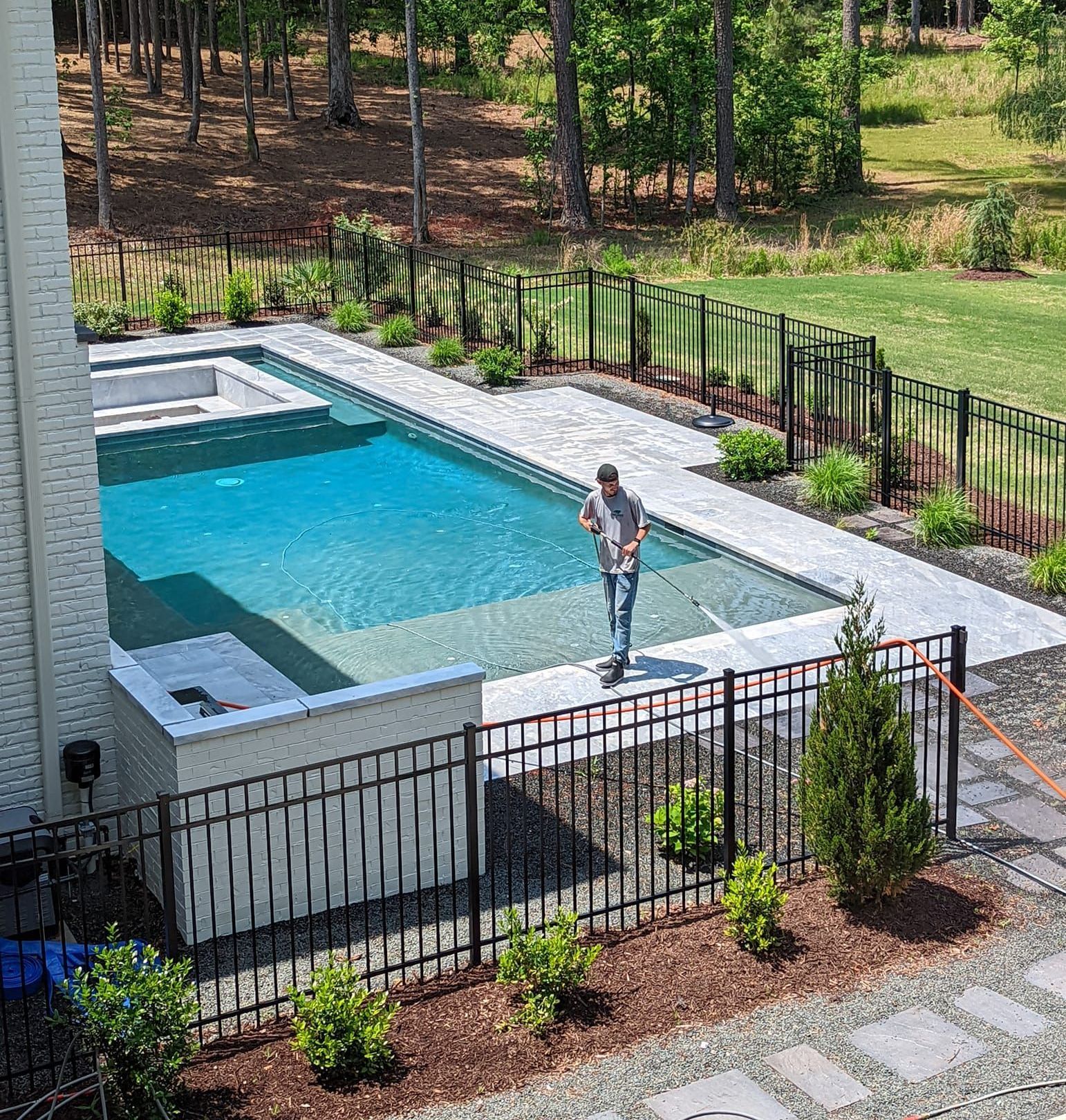 A person stands on the ledge of a rectangular backyard pool with blue water, surrounded by light pavers and a metal fence.