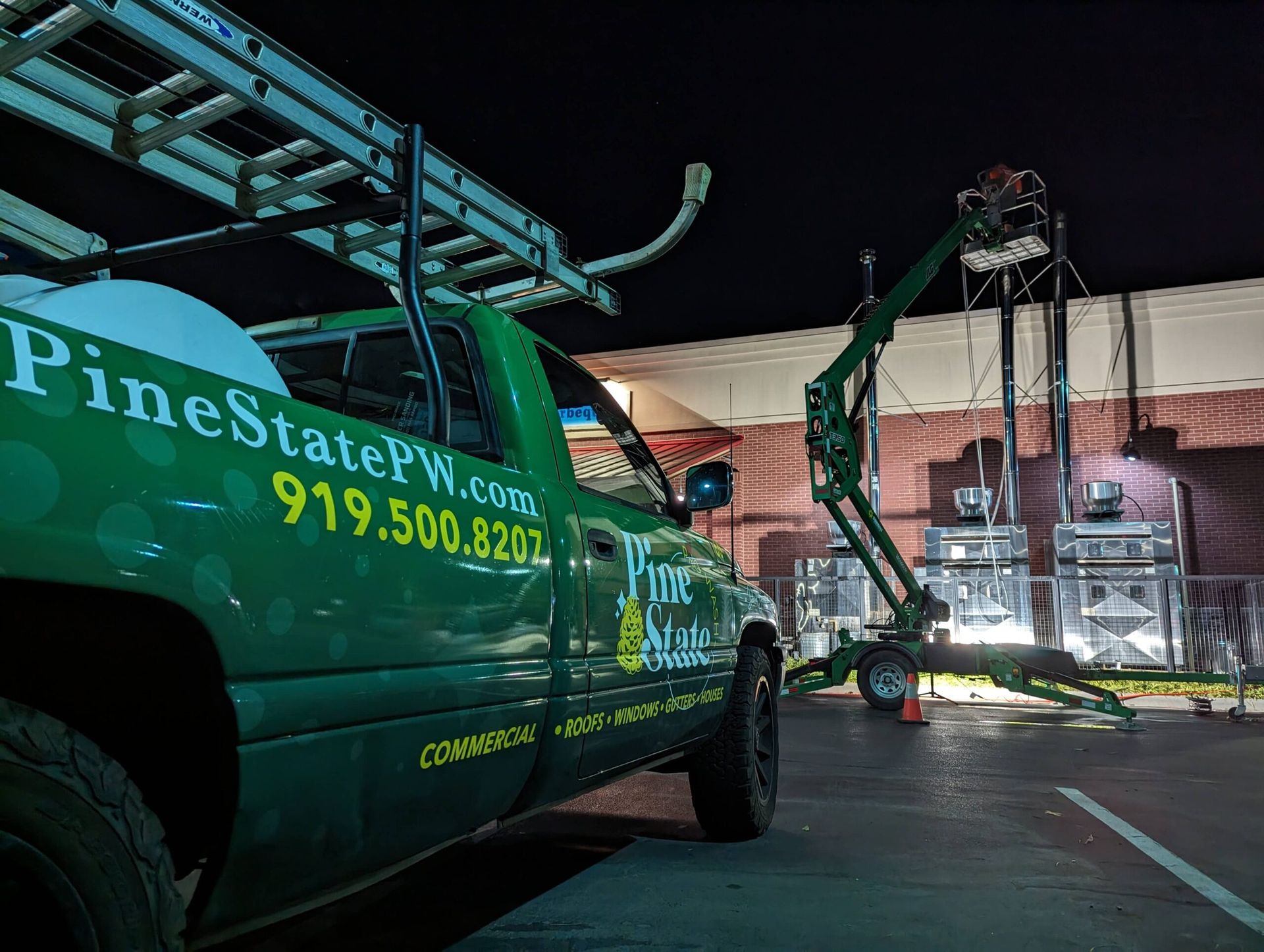 A green Pine State Power Washing truck parked at night next to a mobile lift cleaning the vent hoods of a brick building.