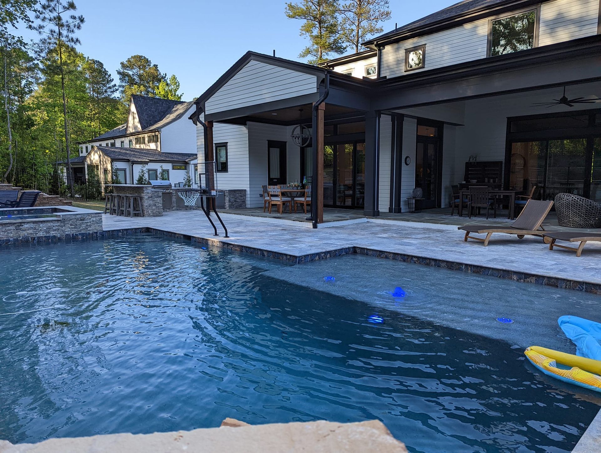 A backyard swimming pool with a stone patio, patio furniture, and a house with a covered porch under a clear blue sky.