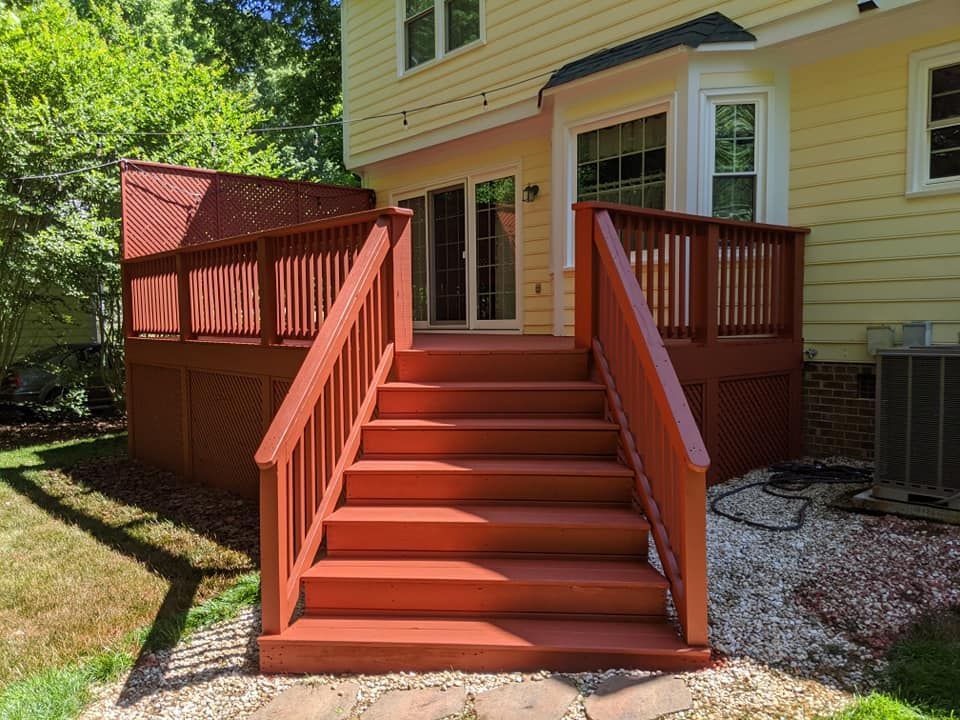 A red wooden deck with stairs leading up to a beige house, surrounded by grass and a gravel yard on a sunny day.