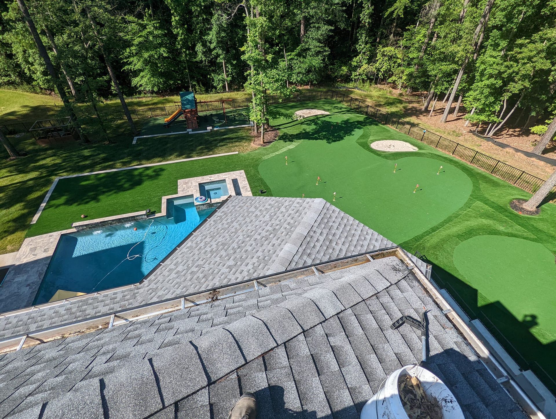 An aerial view of a backyard featuring a swimming pool, a hot tub, and a multi-hole putting green beside a wooded area.