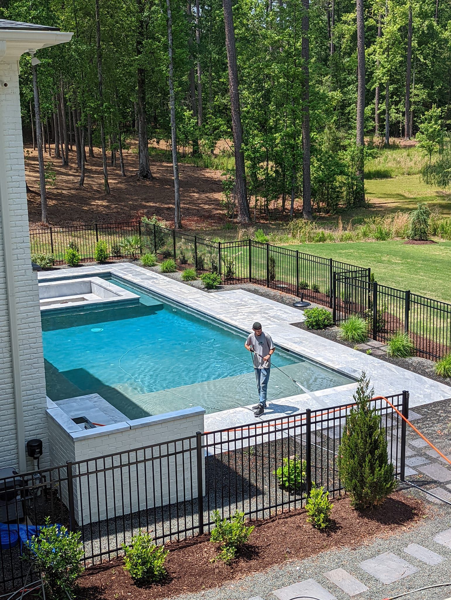 A person stands in a turquoise swimming pool surrounded by a patio and metal fencing, backed by a wooded area.