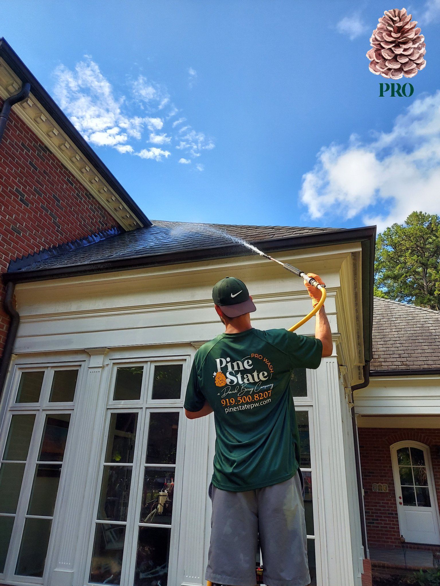 A person wearing a green shirt sprays water from a hose onto a dark roof of a brick house under a blue sky.
