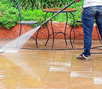 A person in jeans uses a pressure washer to clean a tiled patio near a table and chair set.