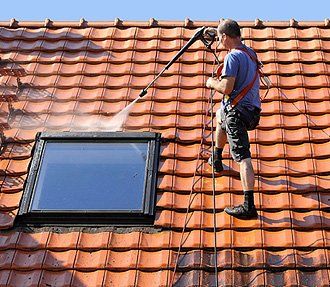A person wearing safety gear uses a power washer to clean orange roof tiles near a skylight window.