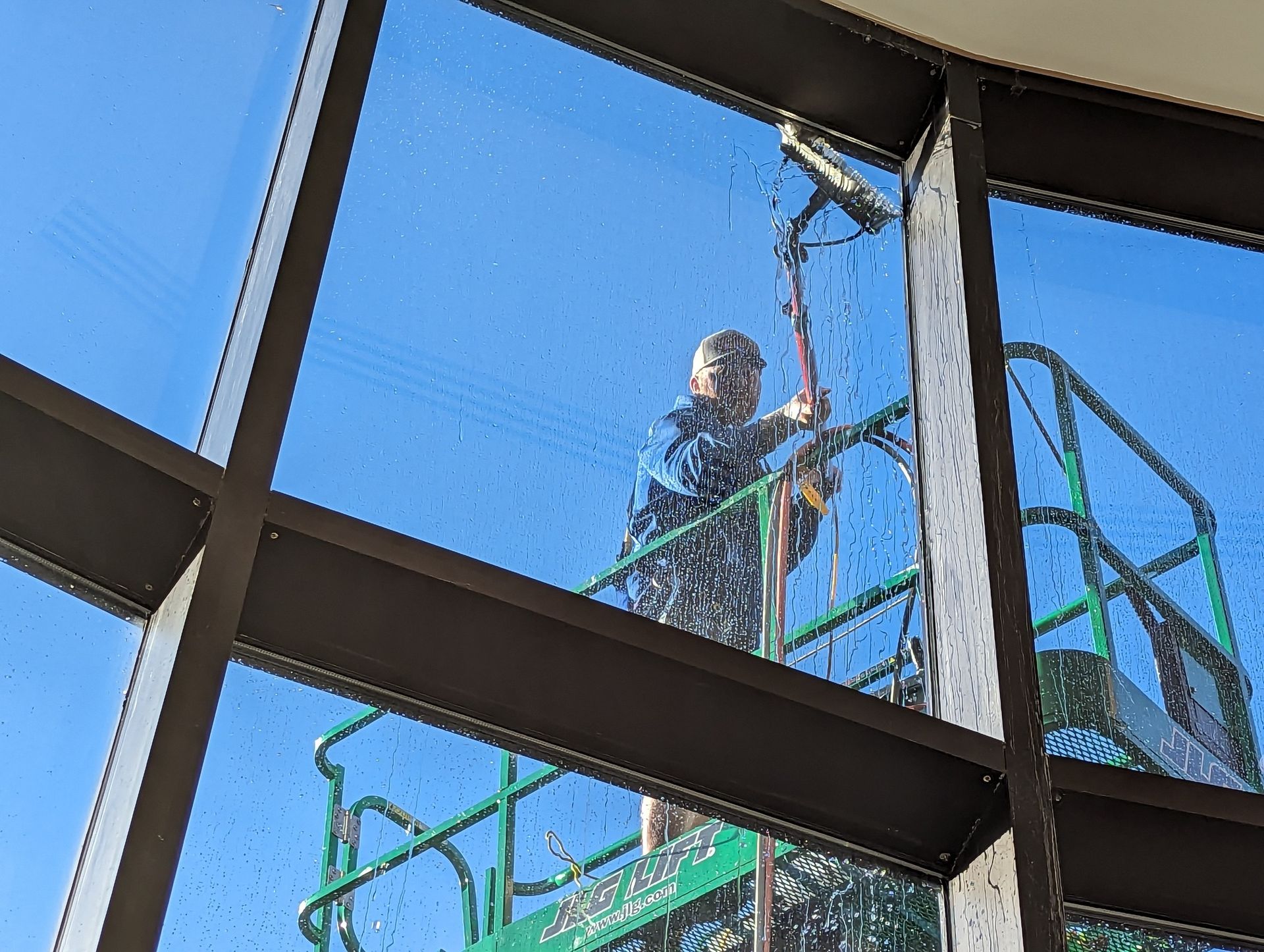 A worker on an elevated platform cleans tall exterior glass windows with a long-handled brush against a blue sky.