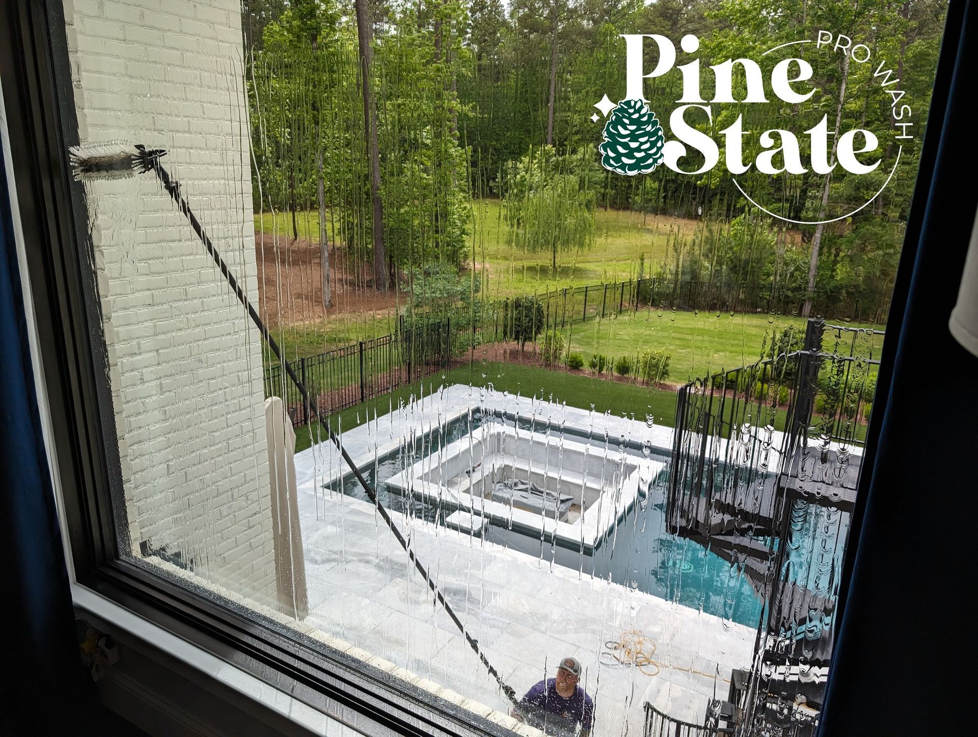 A worker uses a long-reach pole to clean an exterior window above a backyard swimming pool with a central fire pit.