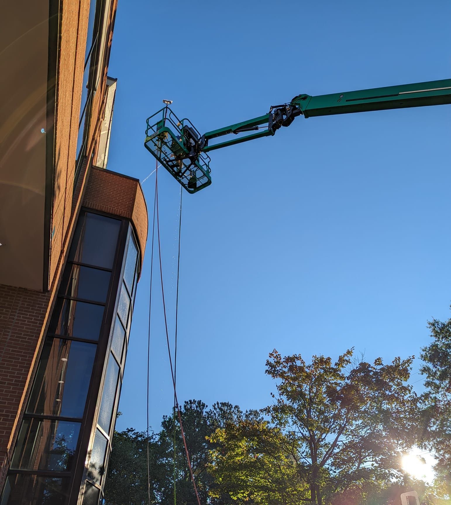A green construction boom lift with a worker in the basket reaches toward the corner of a brick building under a blue sky.