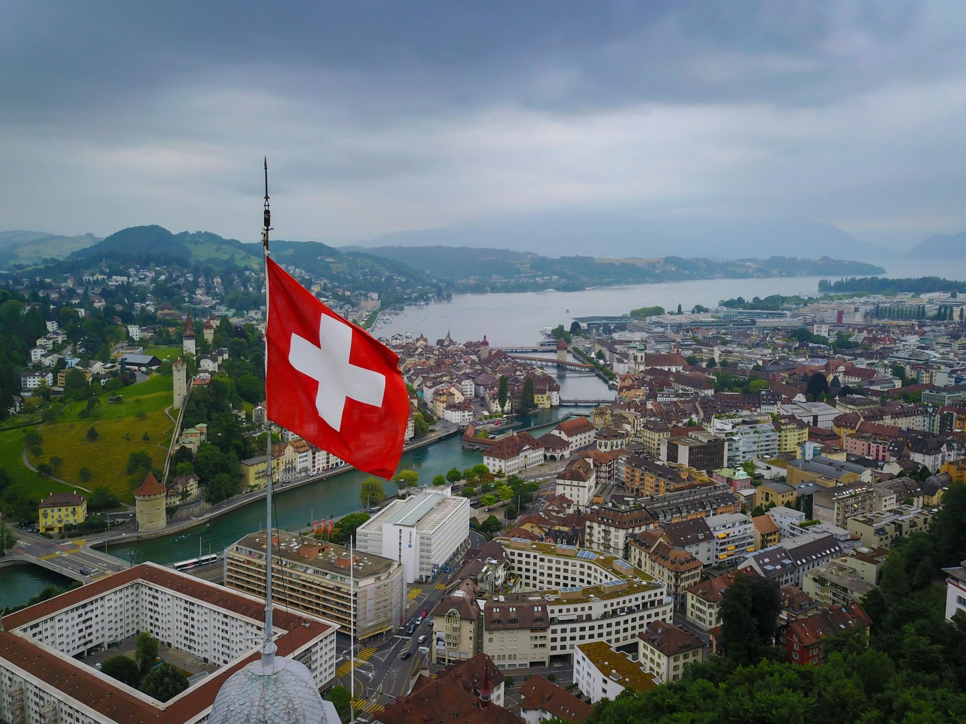 Bandera suiza ondeando sobre una ciudad y un lago en un día nublado.
