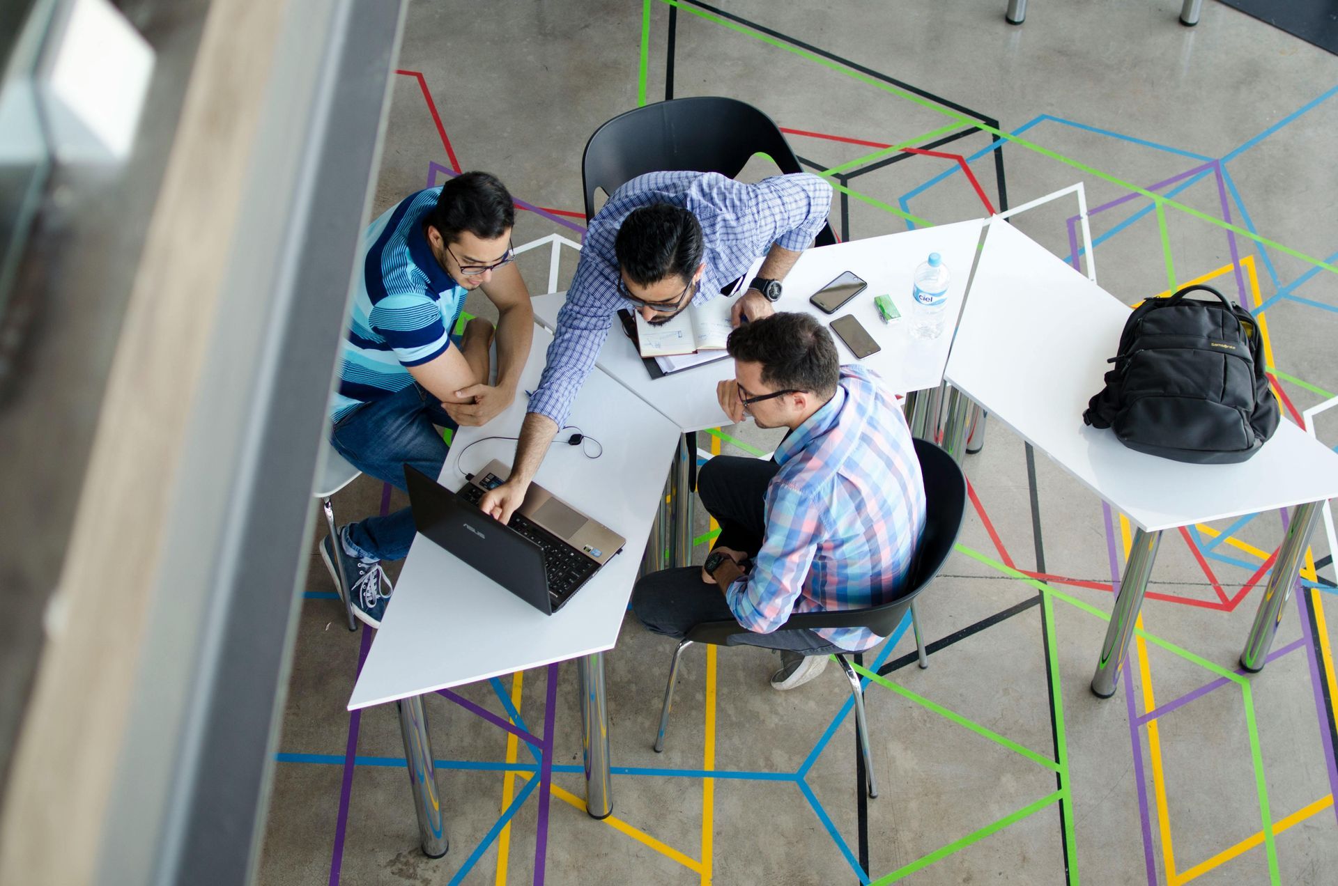 Tres personas alrededor de un escritorio, mirando una computadora portátil. La habitación tiene diseños geométricos en el suelo.