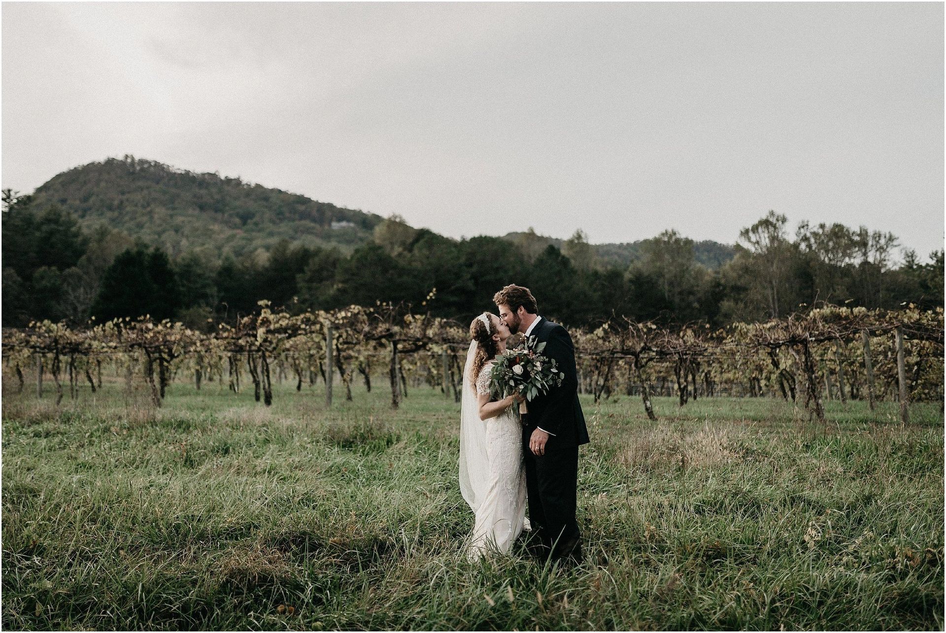 A bride and groom are kissing in a field with mountains in the background.