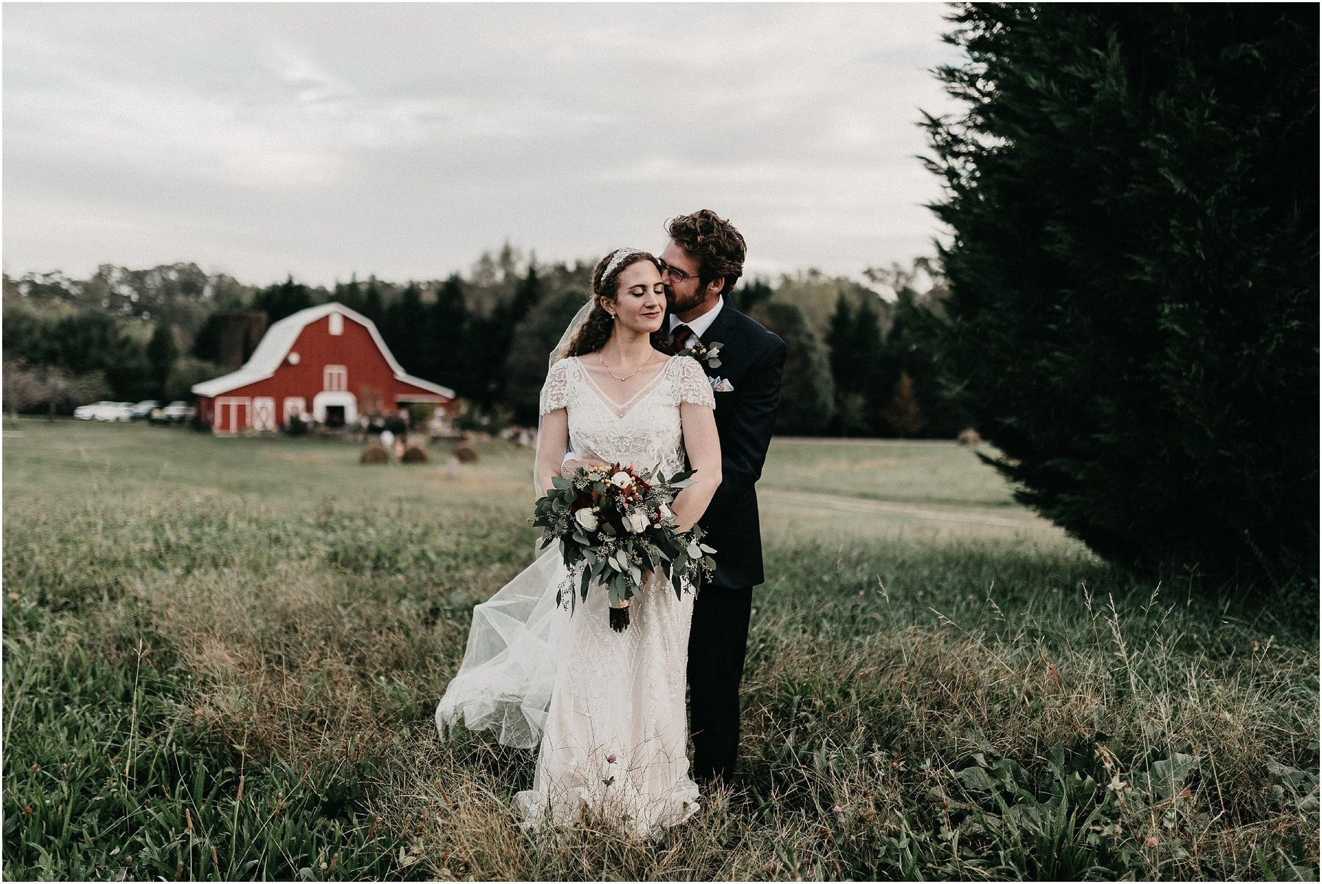 A bride and groom are kissing in a field with a red barn in the background.