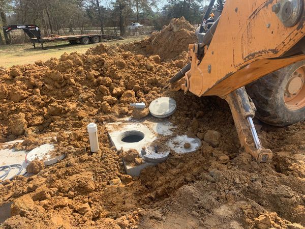 Excavator digging around a septic tank in muddy ground at a rural site