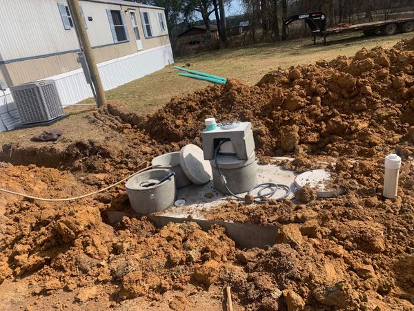 Construction site with dug-up dirt, concrete blocks, and utility pipes beside a house.