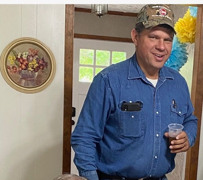 Smiling man in a blue shirt and camo cap holding a drink at a doorway decorated with flowers