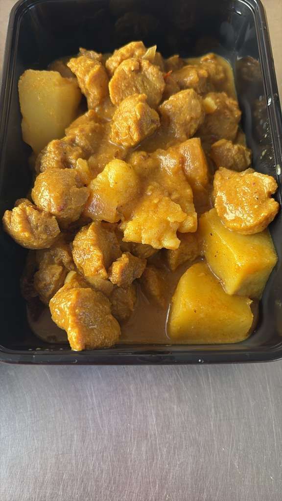 Black takeout container of curry-covered chicken and potatoes on a gray table.