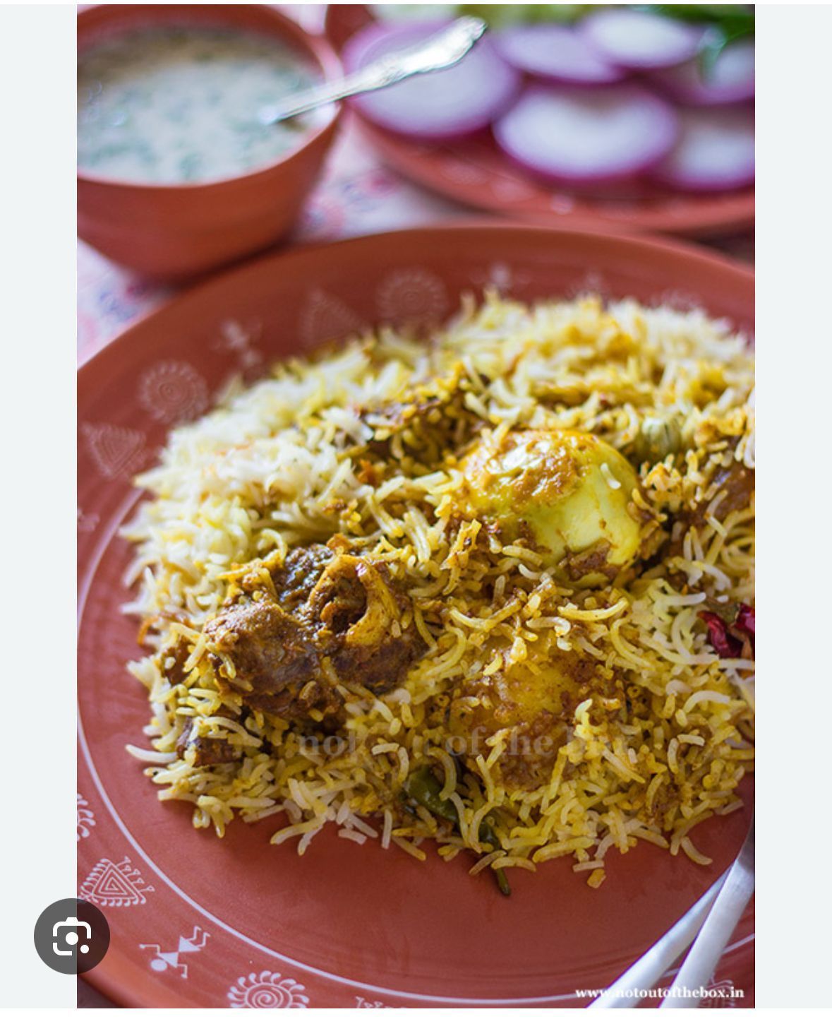 Plate of biryani rice with meat and egg, served with sliced onions and a side of raita.