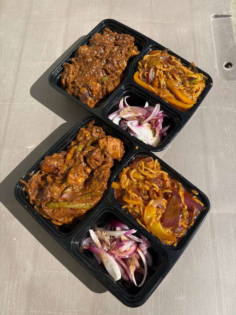 Takeout tray with four saucy meat portions, onions, and fries on a gray table.