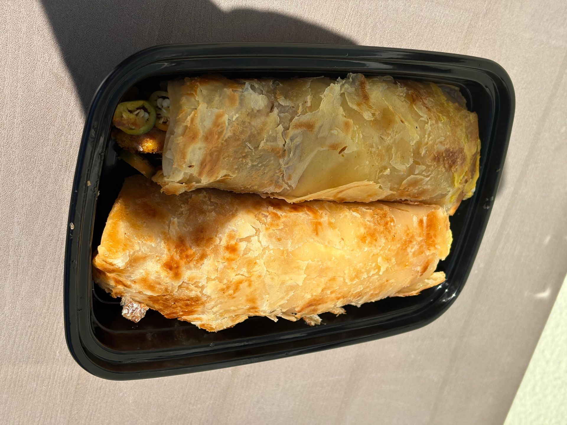 Black takeout container holding two golden-brown wrapped rolls on a light surface