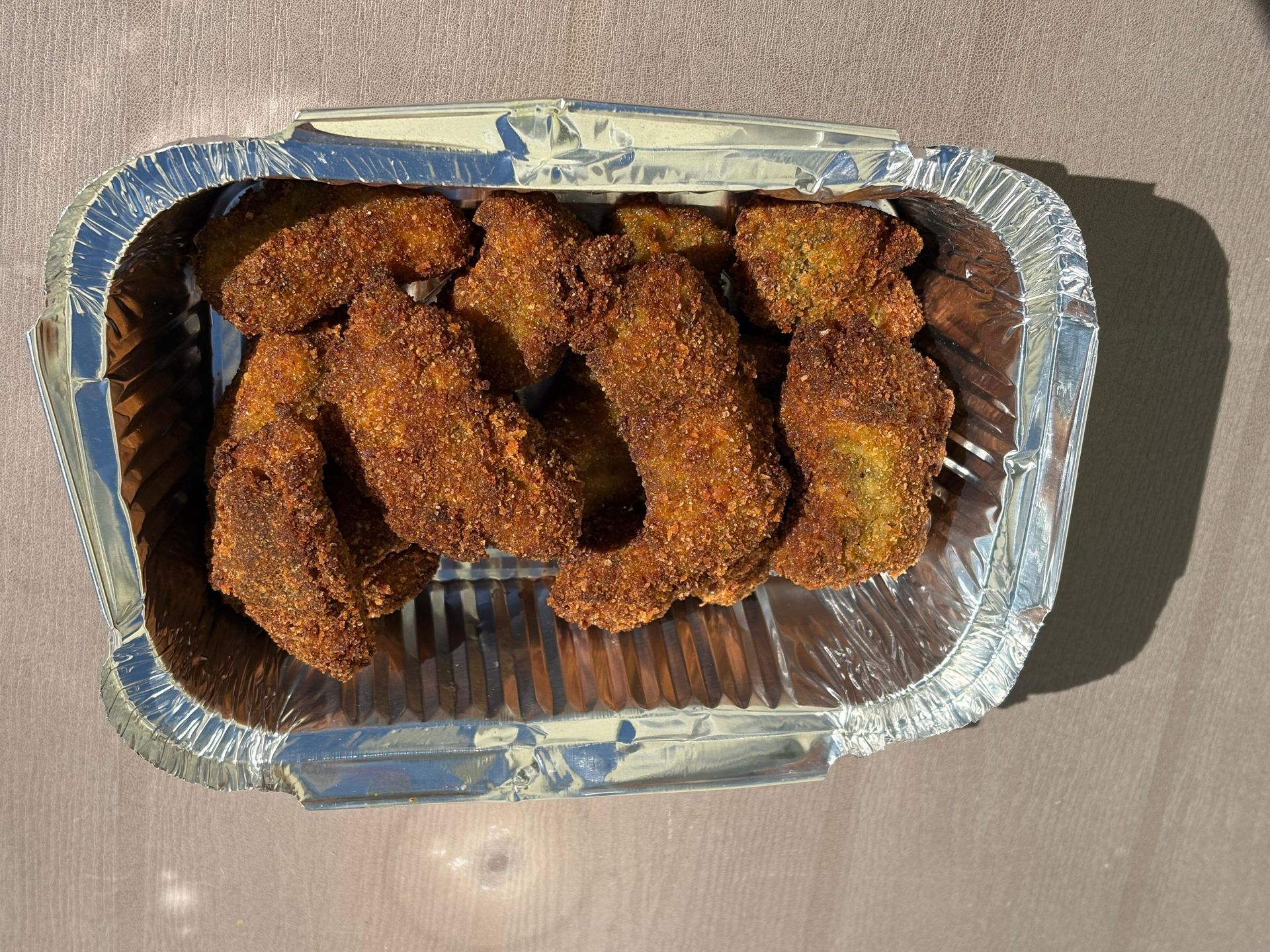 Foil tray of breaded, golden-brown fried food pieces in sunlight