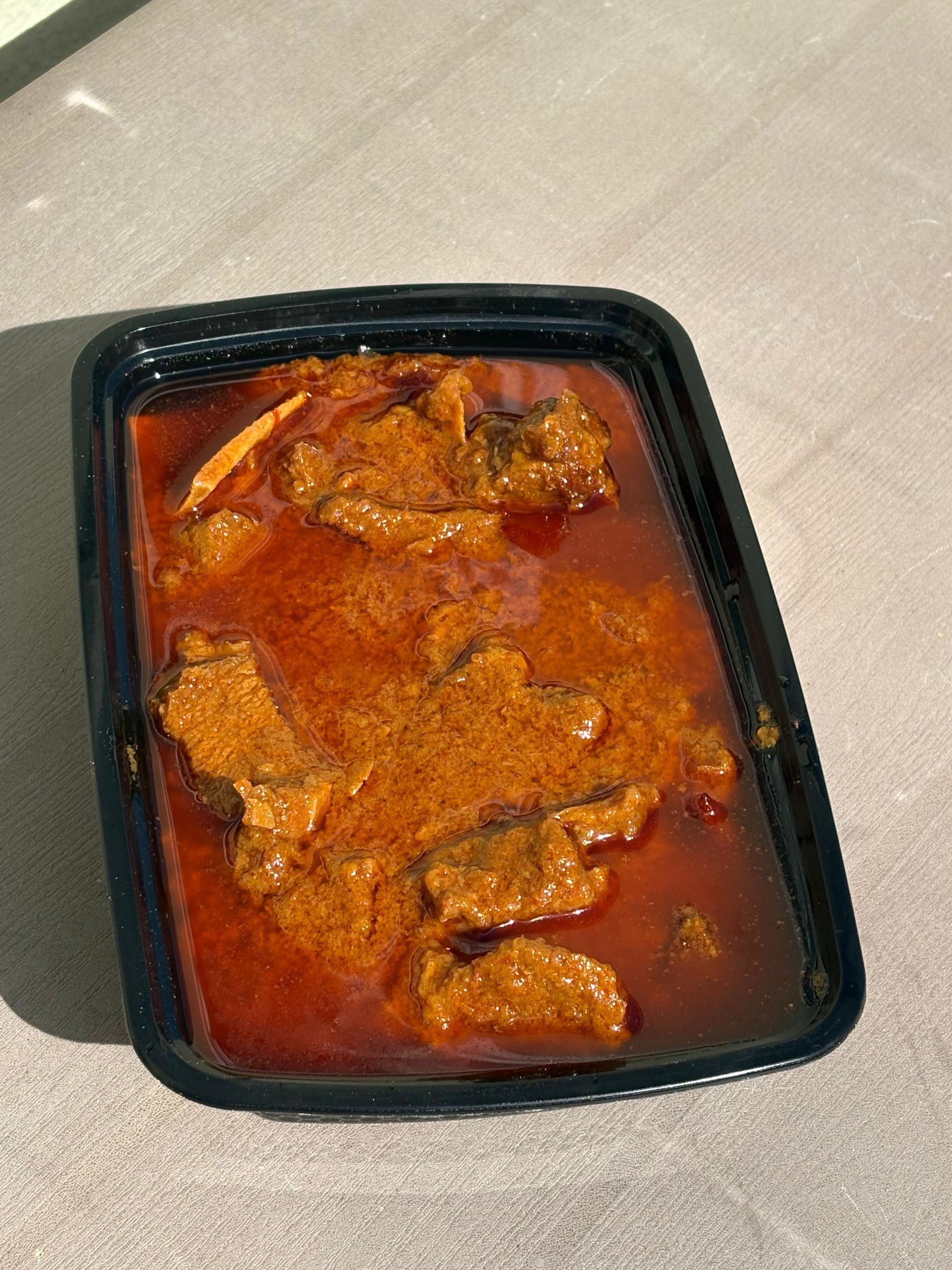 Black container of red curry with meat pieces on a sunlit surface