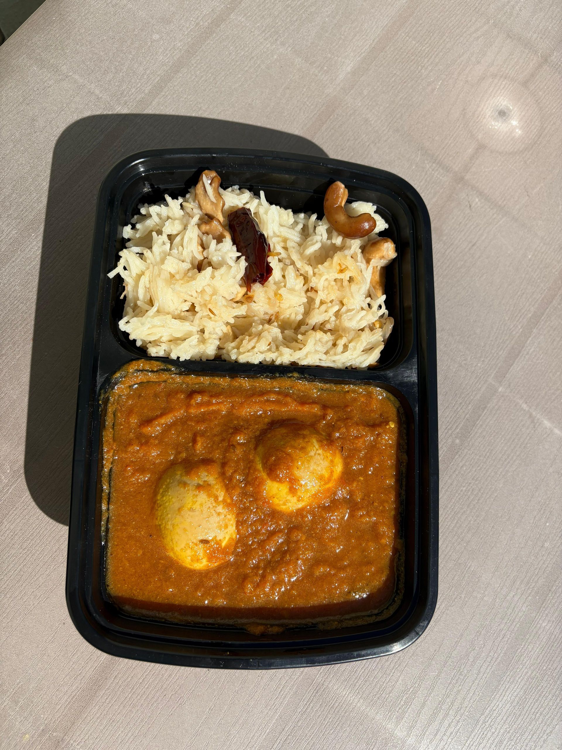 Black takeout tray with white rice and orange curry topped with two boiled eggs.