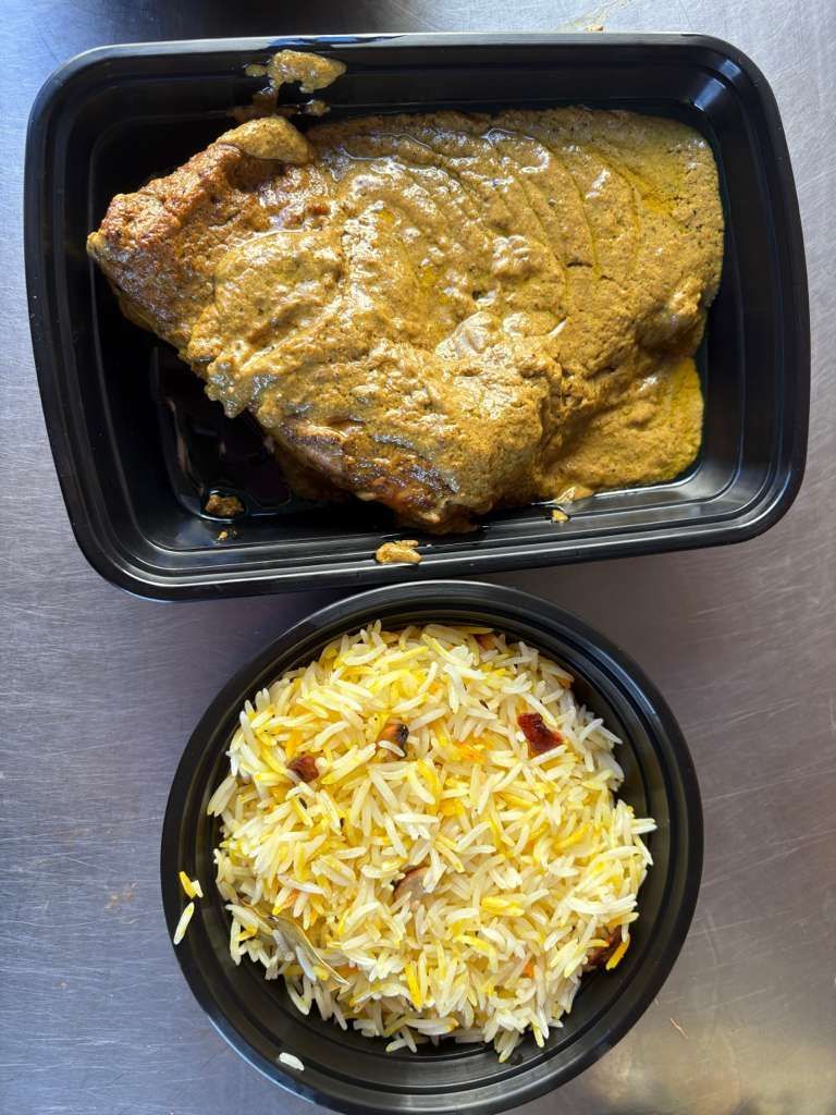 Black takeout tray with curry-covered fried item and a bowl of white rice with yellow seasoning.