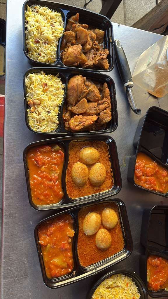 Stacked black meal trays with rice, fried chicken, and orange sauce with round fried snacks on a table.