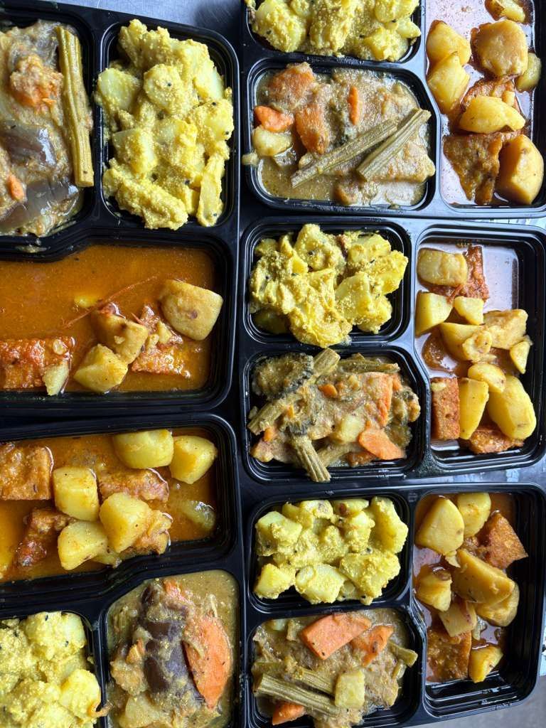 Assorted meals in black takeout trays, including yellow rice, potatoes, curry, and vegetables.