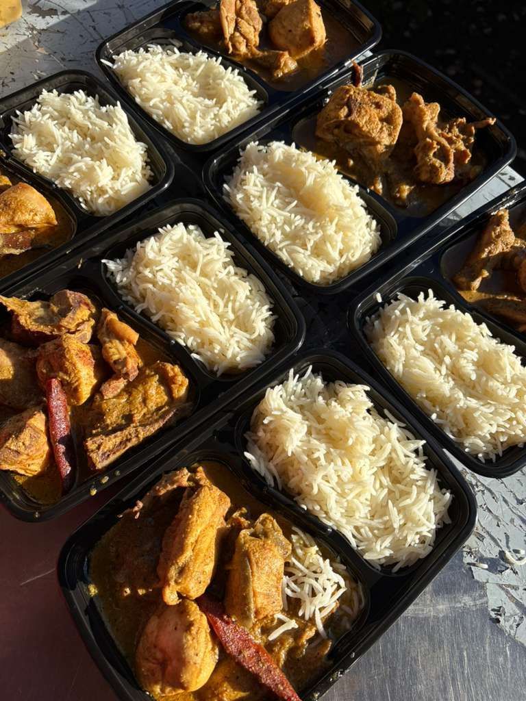 Several black takeout trays with white rice and fried chicken on a sunlit pavement