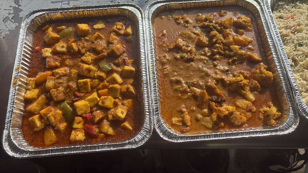 Two foil trays of saucy cooked food with diced vegetables, side by side on a table.
