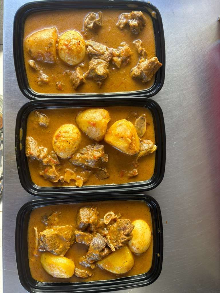 Three black meal trays with curry and potato chunks stacked vertically on a light surface