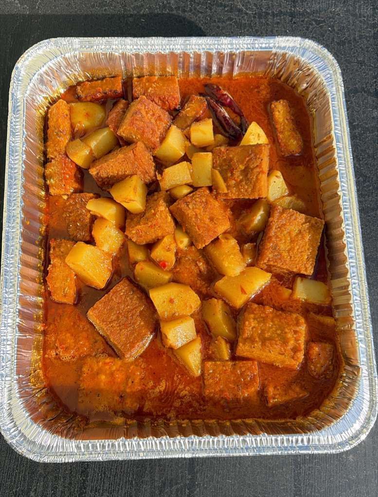 Foil takeout tray of stew with cubed potatoes, carrots, and meat in red broth