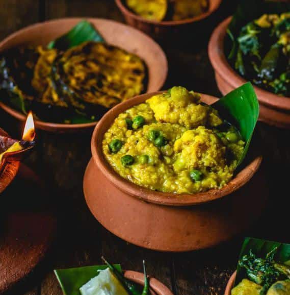 Clay pots of yellow-green curry garnished with herbs on a dark table, with banana leaves visible.