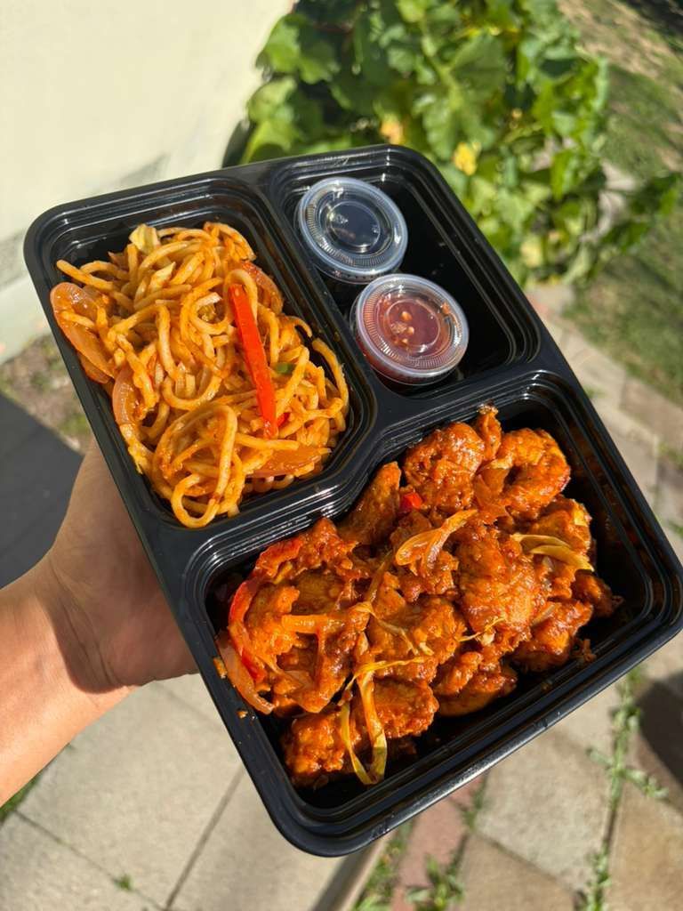 Handheld takeout tray with saucy chicken, noodles, and two dipping cups outdoors