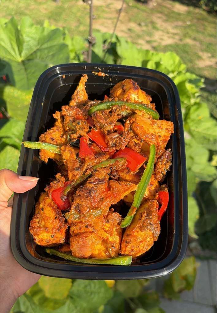 Hand holding a black takeout container of spicy fried tofu with red peppers and green onions outdoors