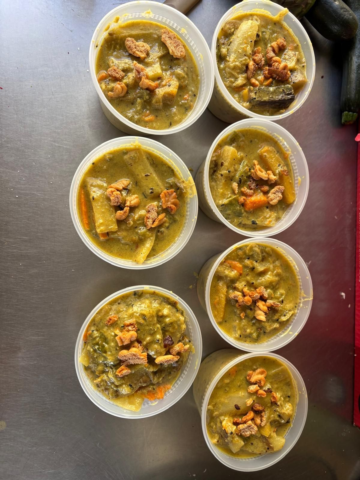 Eight white bowls of vegetable stew on a metal tray, with green broth and chunks of carrots and vegetables.