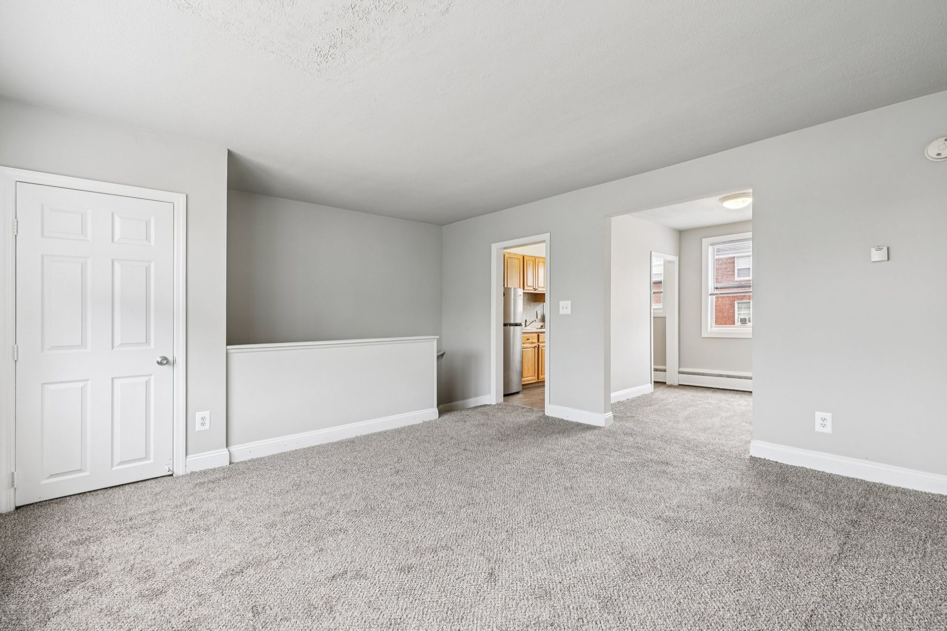 Empty living room with gray carpet, walls, and a white door. A hallway with a kitchen is visible.