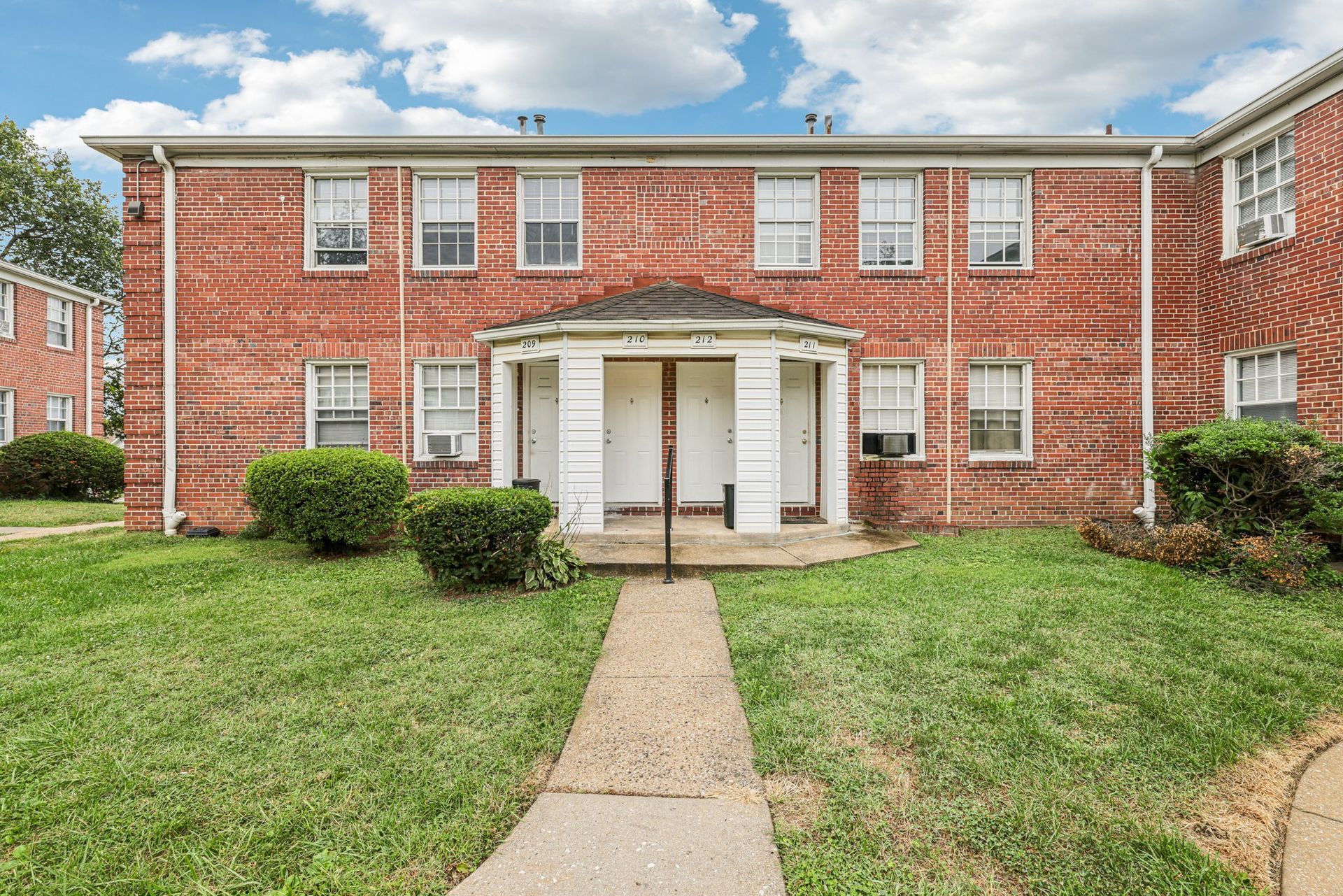 Red brick two-story apartment building with white doors and windows; short bushes and green lawn.